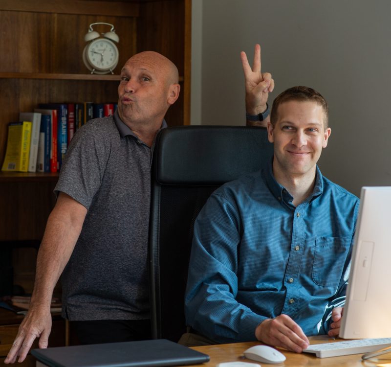 NNPA2024-111 Two men are in a home office setting. One is seated at a desk with a computer, smiling. The other stands behind, making a playful gesture with a peace sign. A bookshelf and clock are in the background.